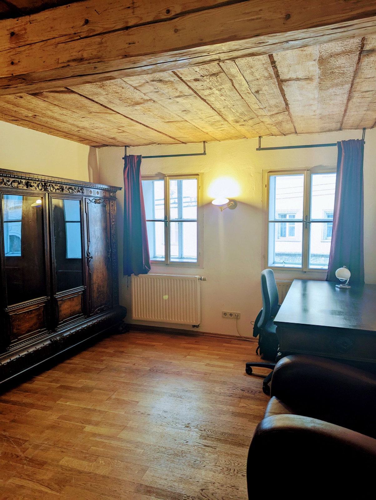 Wohnschlafzimmer mit Holzdecke, Parkettboden, Schreibtisch, altem Holzschrank und zwei Fenstern